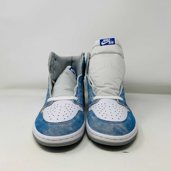 The Jordan 1 Hyper Royal, a high-top sneaker in white and light blue with a gray Nike swoosh and iconic Air Jordan logo, is showcased on a black and red Nike shoebox. It includes extra laces and features a crisp white midsole.
