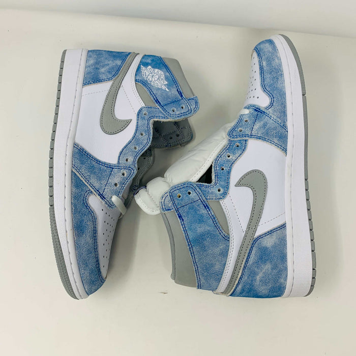 The Jordan 1 Hyper Royal, a high-top sneaker in white and light blue with a gray Nike swoosh and iconic Air Jordan logo, is showcased on a black and red Nike shoebox. It includes extra laces and features a crisp white midsole.