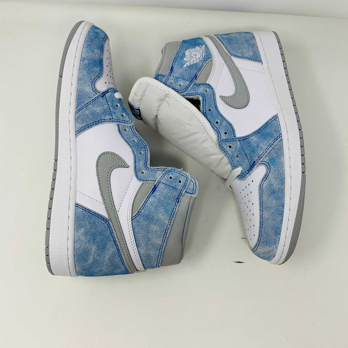 The Jordan 1 Hyper Royal, a high-top sneaker in white and light blue with a gray Nike swoosh and iconic Air Jordan logo, is showcased on a black and red Nike shoebox. It includes extra laces and features a crisp white midsole.