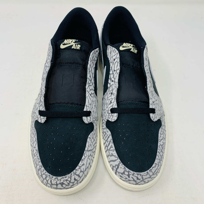 The Jordan 1 Retro Low OG Black Cement sneaker, with black and gray elephant print and a white swoosh, is showcased on a matching shoe box. A small brown box with extra laces is beside it, all set against a plain background.