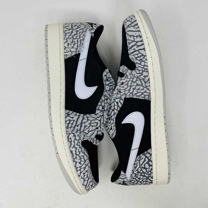 The Jordan 1 Retro Low OG Black Cement sneaker, with black and gray elephant print and a white swoosh, is showcased on a matching shoe box. A small brown box with extra laces is beside it, all set against a plain background.