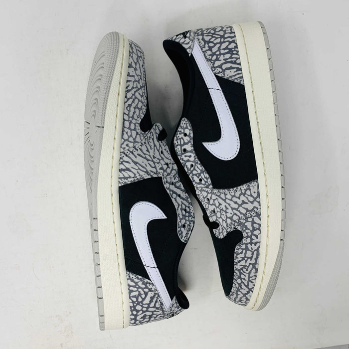 The Jordan 1 Retro Low OG Black Cement sneaker, with black and gray elephant print and a white swoosh, is showcased on a matching shoe box. A small brown box with extra laces is beside it, all set against a plain background.