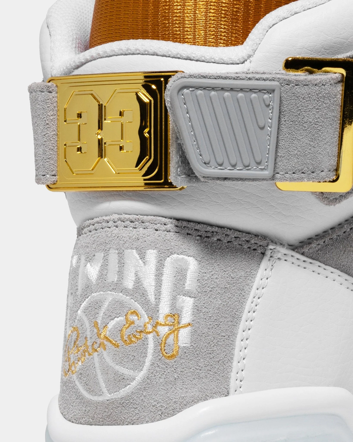 Ewing Athletics 33 HI x LAURENS J sneakers close-up showing gray suede panels, white leather, embroidered logos, and gold metallic "33" strap detail.