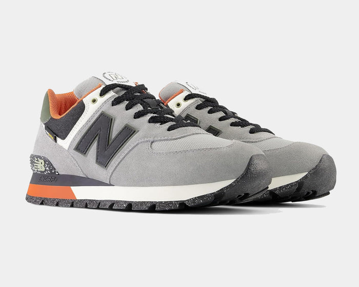 New Balance 574 Rugged sneakers in gray suede with black details, orange accents, and speckled outsole, angled front view.