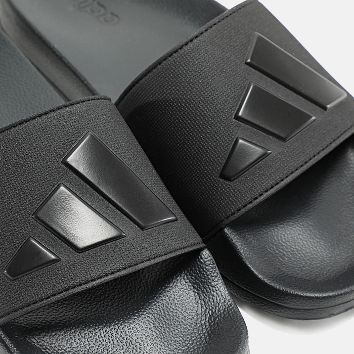 Adidas Adilette Shower Slides in black with textured footbed, synthetic strap, and embossed logo detail, close-up view.