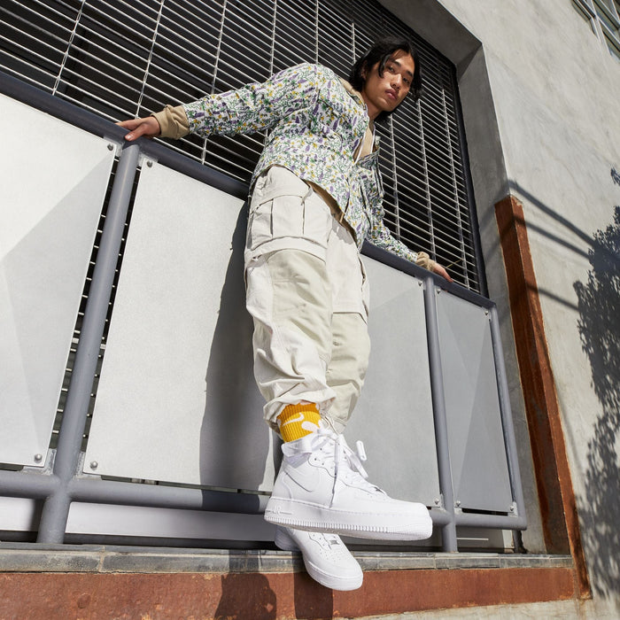 Nike Air Force 1 Mid '07 sneakers in white leather with perforated toe box and ankle strap, worn by person leaning on a railing outdoors.