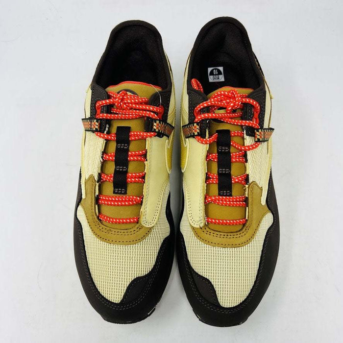 A brand new Nike Air Max 1 TS Cactus Jack Baroque Brown sneaker in yellow and brown with orange accents sits on a brown Nike shoebox. It boasts a wavy design, textured fabric, extra brown laces, and a visible air unit in the sole against a plain white background.