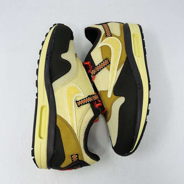 A brand new Nike Air Max 1 TS Cactus Jack Baroque Brown sneaker in yellow and brown with orange accents sits on a brown Nike shoebox. It boasts a wavy design, textured fabric, extra brown laces, and a visible air unit in the sole against a plain white background.