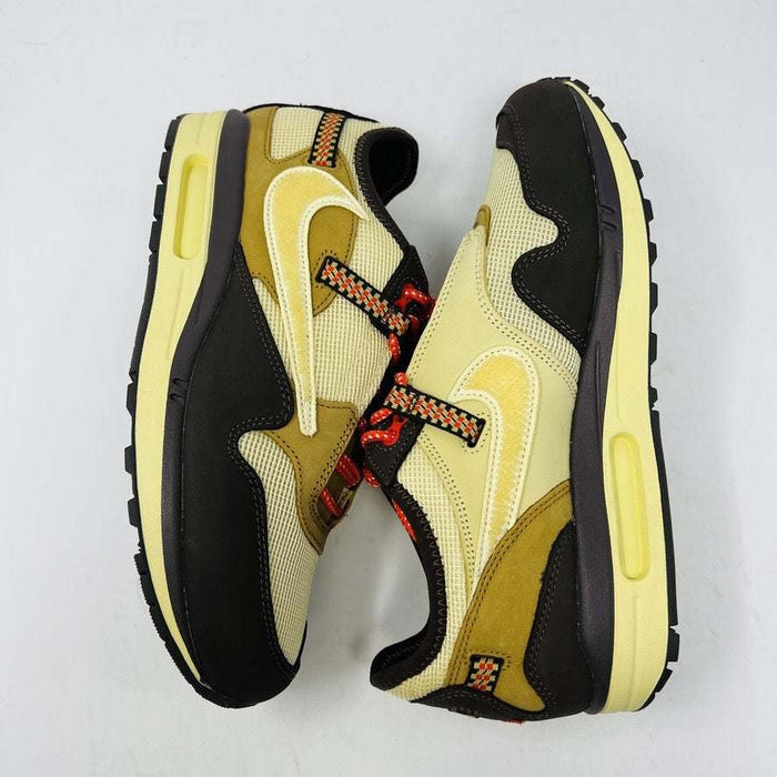 A brand new Nike Air Max 1 TS Cactus Jack Baroque Brown sneaker in yellow and brown with orange accents sits on a brown Nike shoebox. It boasts a wavy design, textured fabric, extra brown laces, and a visible air unit in the sole against a plain white background.
