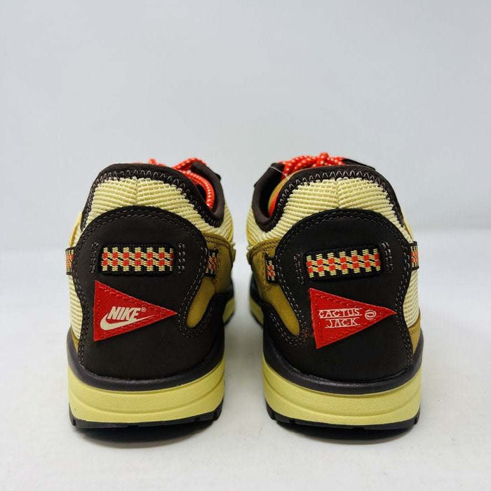 A brand new Nike Air Max 1 TS Cactus Jack Baroque Brown sneaker in yellow and brown with orange accents sits on a brown Nike shoebox. It boasts a wavy design, textured fabric, extra brown laces, and a visible air unit in the sole against a plain white background.