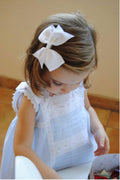Child wearing a white lace dress with a large bow in their hair, sitting on a tiled floor.