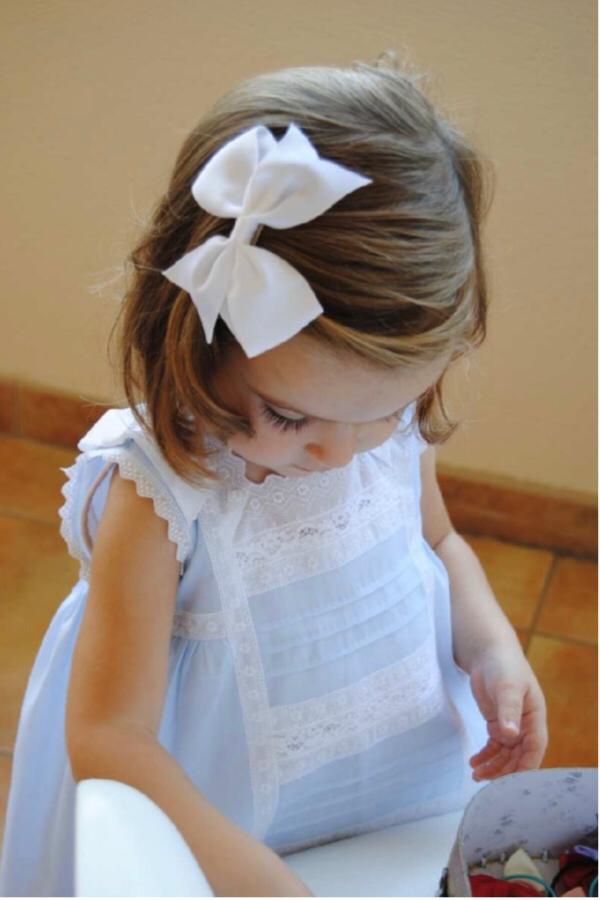 Child wearing a white lace dress with a large bow in their hair, sitting on a tiled floor.