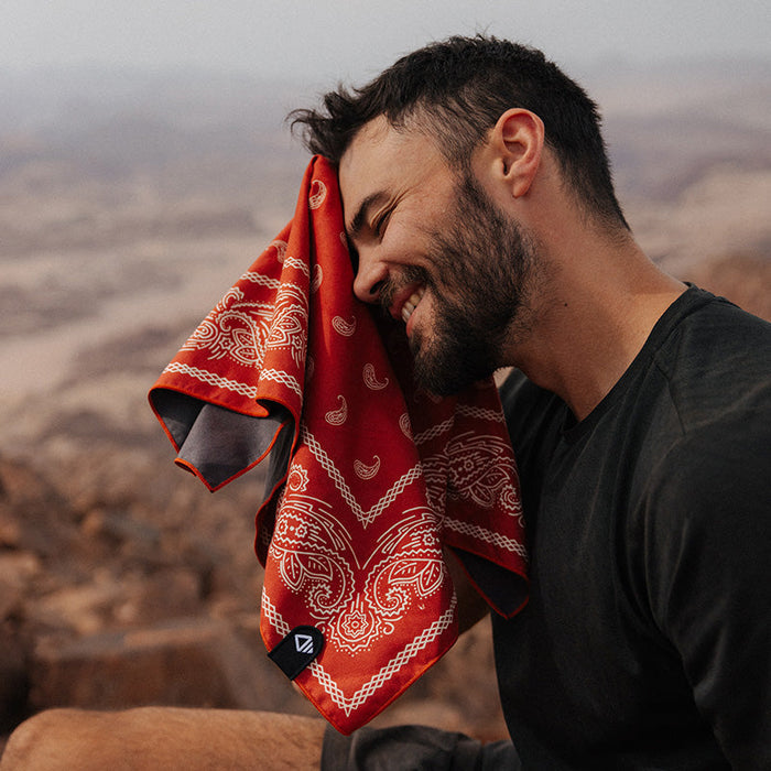 Nomadix Bandana Towel in red with paisley design and black trim, lightweight and soft material, outdoor lifestyle setting.