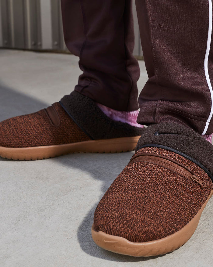 Nike Burrow SE slippers in brown with textured knit upper, zippered top detail, fleece lining, and rubber outsole, lifestyle context