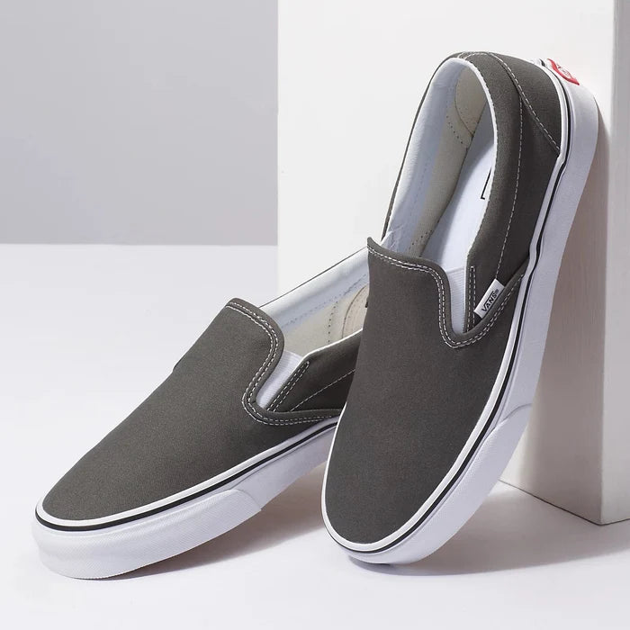 Vans Classic Slip On shoes in dark gray canvas with white rubber sole and elastic side accents, angled pair view.