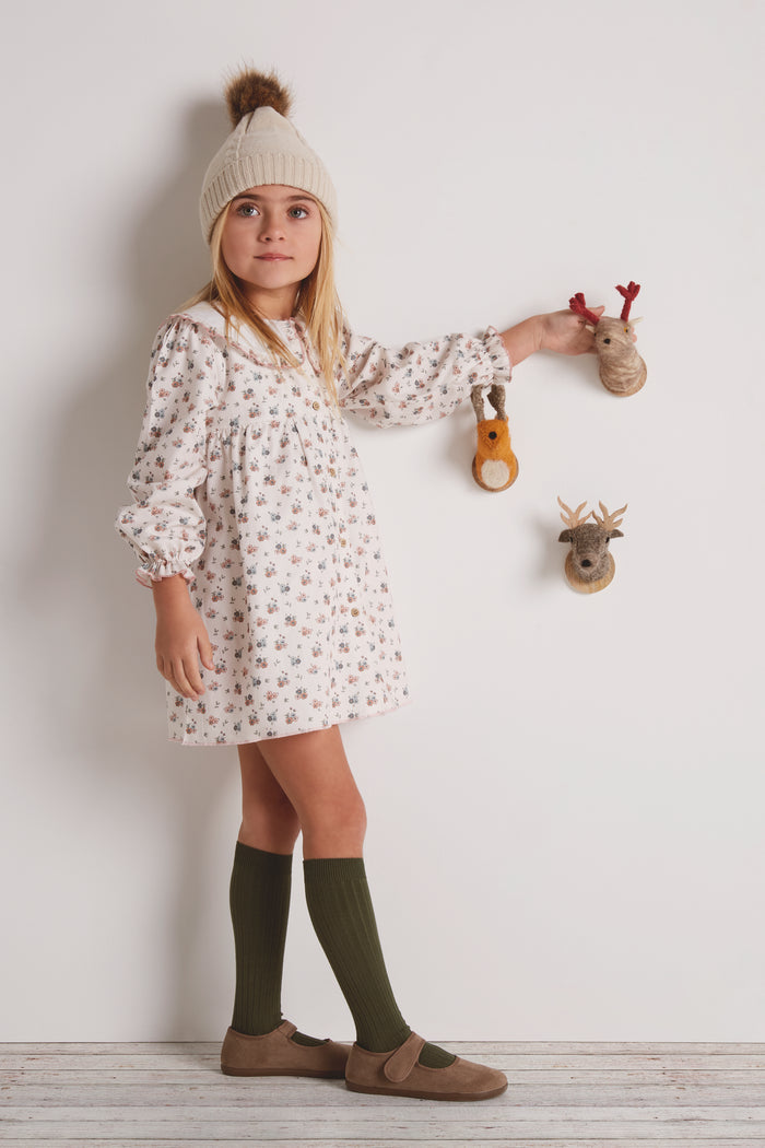 Young girl in a floral dress with green socks and brown shoes standing against a white wall.