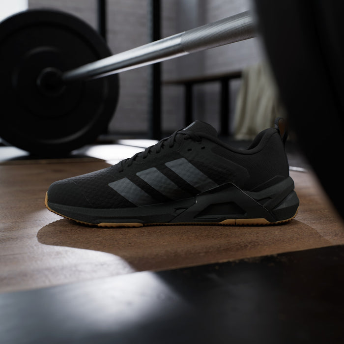 Adidas Dropset Control Trainer shoes in black with textured upper, gray stripes, and gum sole, side view placed in a gym setting.