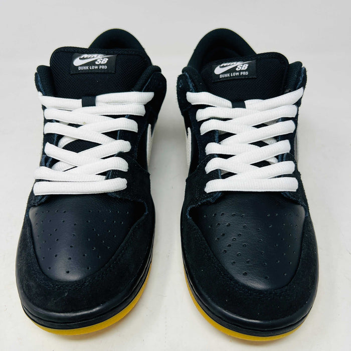 A black Nike Dunk Low Pro SB Black White Gum sneaker with white laces and swoosh sits on a purple Nike SB box. The 2025 shoe has a yellow sole, leather-texture finish, and both are in new condition.