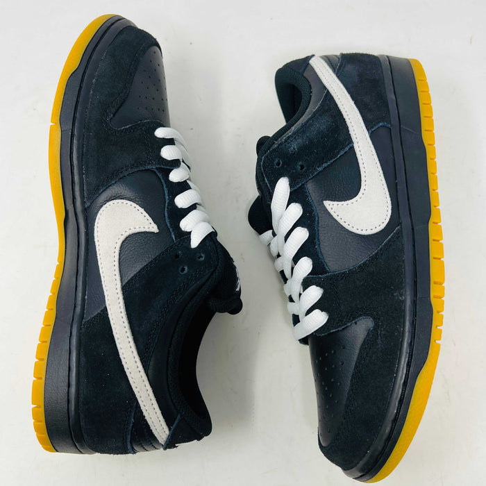A black Nike Dunk Low Pro SB Black White Gum sneaker with white laces and swoosh sits on a purple Nike SB box. The 2025 shoe has a yellow sole, leather-texture finish, and both are in new condition.