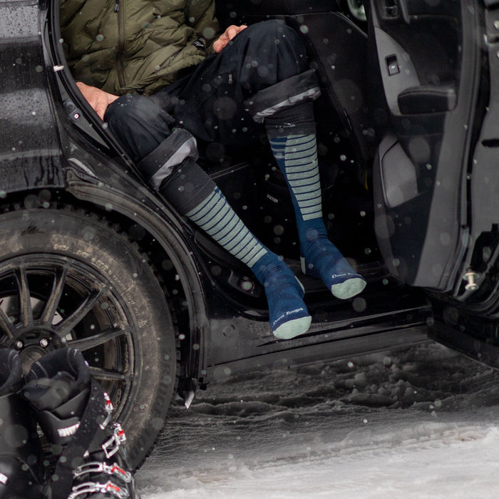 Darn Tough Edge Over-the-Calf Midweight Ski & Snowboard Socks in blue and green stripes with reinforced toe and heel, worn by skier sitting in snowy outdoor setting.