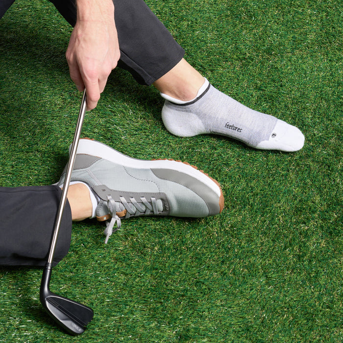 Feetures Elite Golf Max Cushion No Show Tab socks in gray and white, worn by golfer on grass.