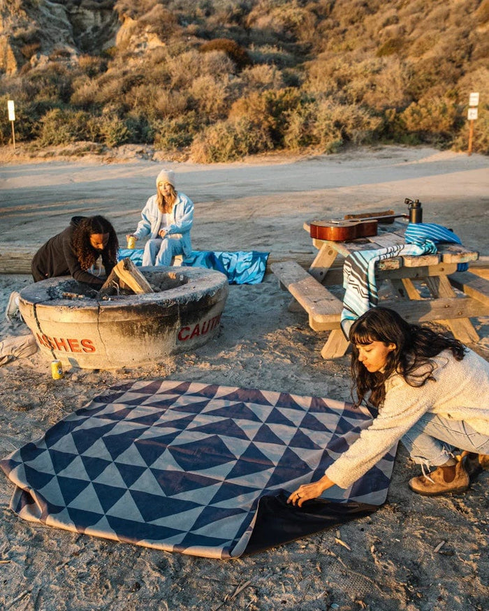 Nomadix Festival Blanket with geometric pattern in navy and gray, spread out on sandy ground near a fire pit and picnic table in an outdoor setting.