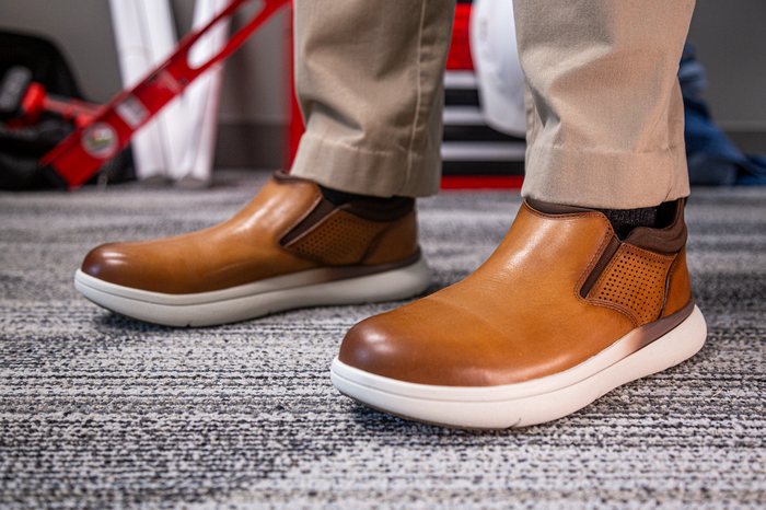 Florsheim Work Fleet Work Comp Toe Slip On work shoes in tan leather with brown elastic gussets, perforated sides, and white slip-resistant soles, worn in workplace setting.