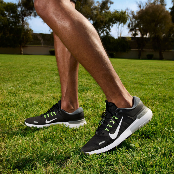 Nike Free Golf Next Nature golf shoes in black mesh with white sole and green lace details, worn by person walking on grass.