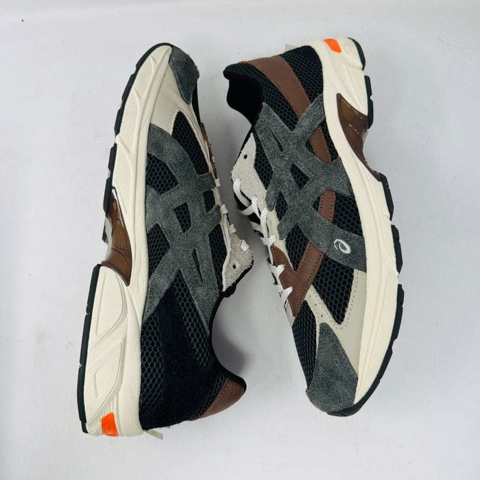 A sleek ASICS Gel-1130 MK-II HAL Studio Forest sits on its box, displaying black and brown mesh with suede accents. Distinctive orange detailing highlights the heel, while an extra brown laces bag accompanies it, enhancing its authentic athletic style.