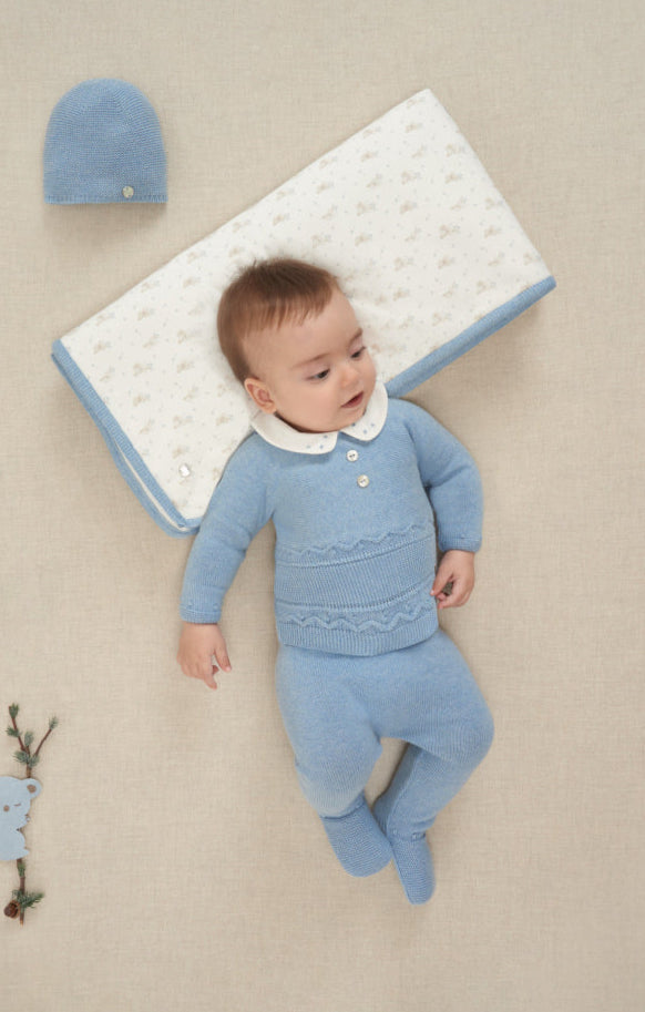 Baby in blue outfit lying on a beige surface with matching hat and blanket.