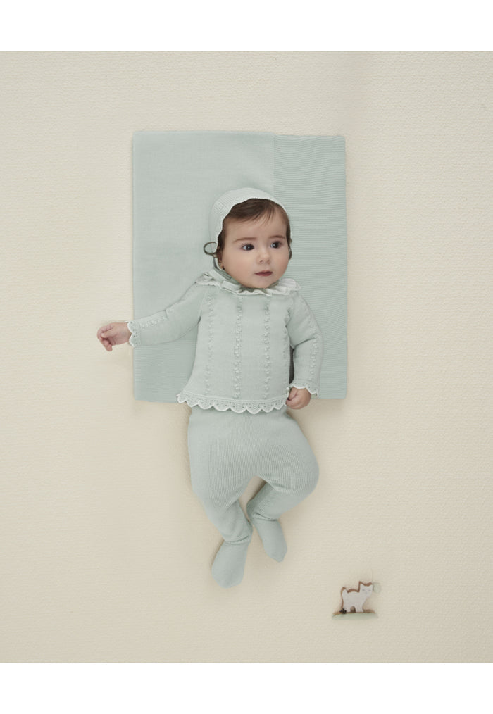 Baby in a light green outfit standing on a white surface with a light blue blanket behind.