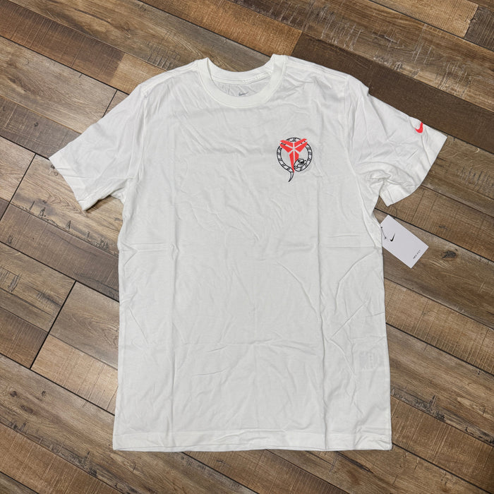 The Nike Kobe Dri-FIT Tee Stockings White features a heart-shaped snake and wings graphic on the left chest. A Nike logo adorns the left sleeve, with a tag attached, all displayed on a wooden floor.