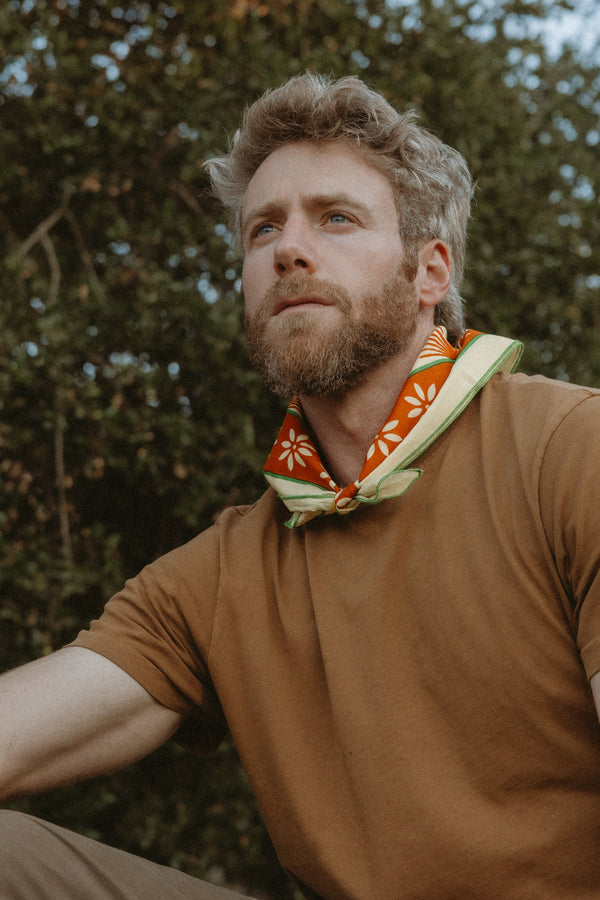 Bandits "La Magia De Tochtli" bandana in orange with white floral design and green border, worn around the neck in an outdoor setting.