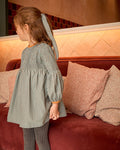 Child in a patterned dress standing next to a red couch with pillows.