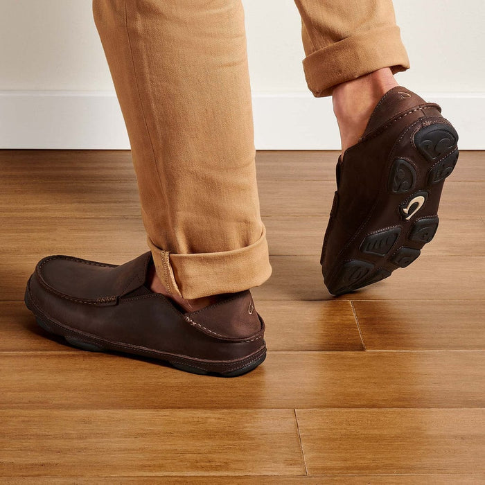 OluKai Moloa shoes in dark brown leather with a collapsible heel design and rubber outsole, lifestyle view worn indoors.