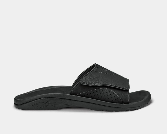 OluKai Nalu Slide sandals in black with textured strap, perforated detailing, and cushioned sole, side view.