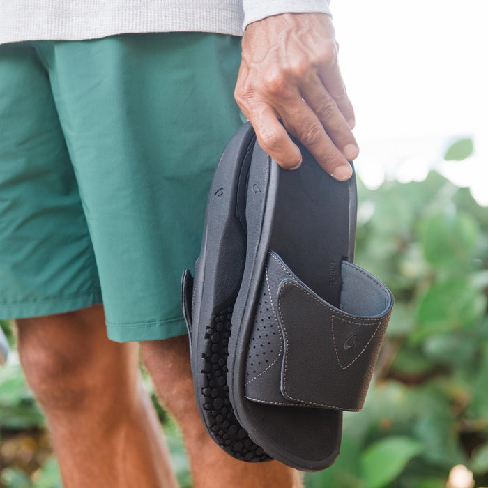 OluKai Nalu Slide sandals in black with perforated strap design and textured outsole, held by person outdoors.