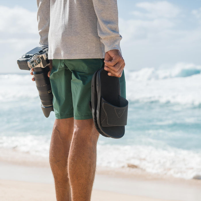 OluKai Nalu Slide sandals in black with adjustable strap and textured footbed, lifestyle view at the beach.