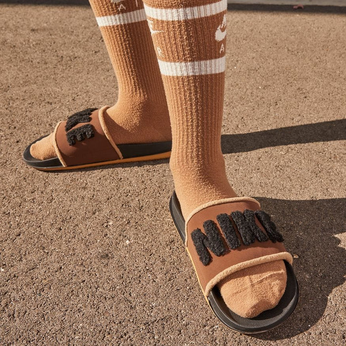 Nike Offcourt Slide SE sandals in brown with plush "NIKE" text detail on strap, worn outdoors with socks.