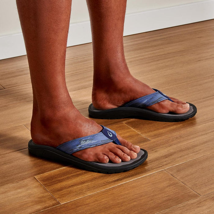 OluKai Ohana Pa'i sandals with blue straps and black cushioned sole, worn by person indoors on wooden flooring.