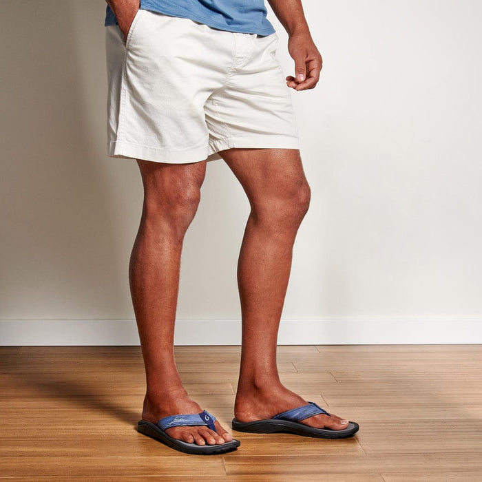 OluKai Ohana Pa'i flip-flops in blue with a cushioned footbed and durable outsole, shown worn indoors on wooden flooring.