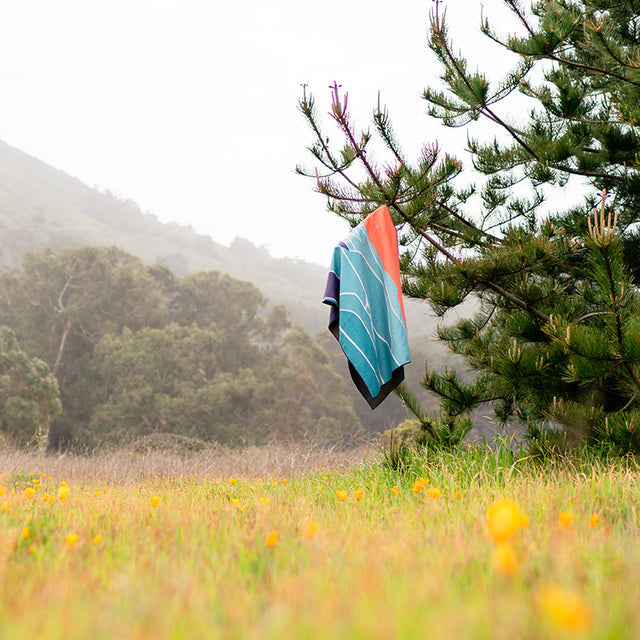 Nomadix Original Towel Big Sur in vibrant teal, orange, and black with wave-inspired design, hanging outdoors in nature.
