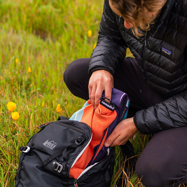 Nomadix Original Towel Big Sur in orange, purple, and teal with a lightweight, packable design, shown being packed into a backpack outdoors.