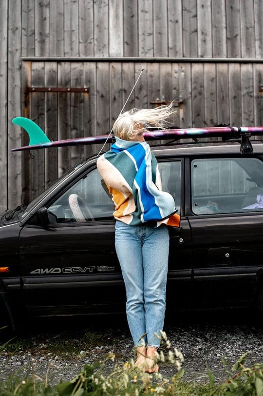 "Nomadix Original Towel in colorful striped pattern featuring blue, orange, and white, worn outdoors by woman near car and surfboard"