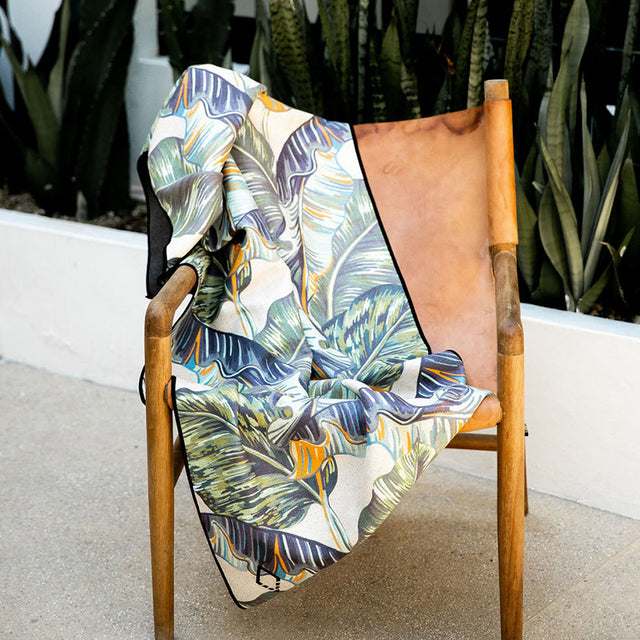 Nomadix Original Towel with tropical leaf pattern in green, orange, and blue hues, draped over a wooden chair outdoors.