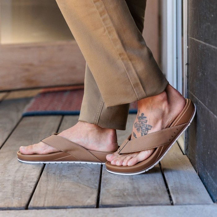 Reef Phantom Bronzer sandals with brown synthetic straps and cushioned footbed, worn by person standing indoors.