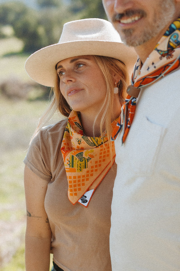 Bandits "Prickly Pollinators" bandana in vibrant orange with cactus and bee illustrations, worn around the neck, outdoor setting.