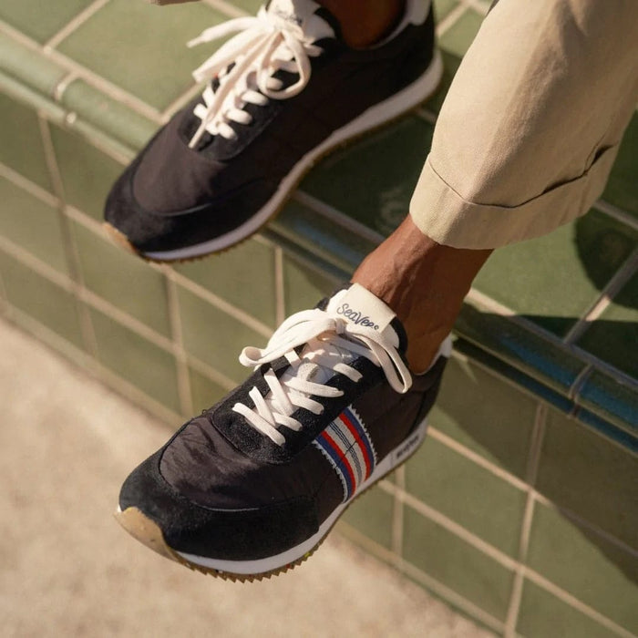 SeaVees Royal Runner sneakers in black with white laces, red and blue striped side details, worn casually outdoors.