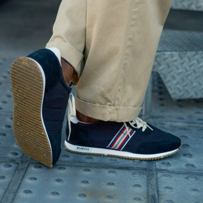 SeaVees Royal Runner sneakers in navy blue with suede panels, striped side accents, and gum rubber outsole, lifestyle view worn on a sidewalk.