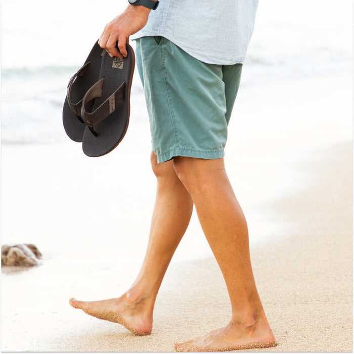 Reef Santa Anna sandals in brown with durable straps and textured footbed, held by a person walking barefoot on a sandy beach.